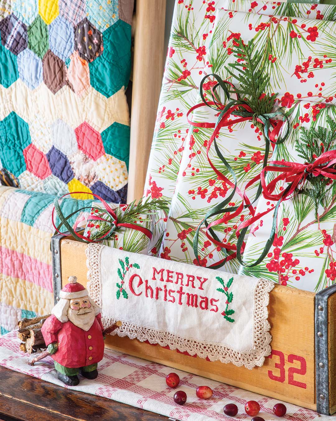 Wrapped gifts nestled within a warm wooden crate are embellished with holly and sprigs of greenery, imparting an earthy charm to the space, and an embroidered handkerchief featuring a classic holiday motif adds another element of heartfelt craftmanship.