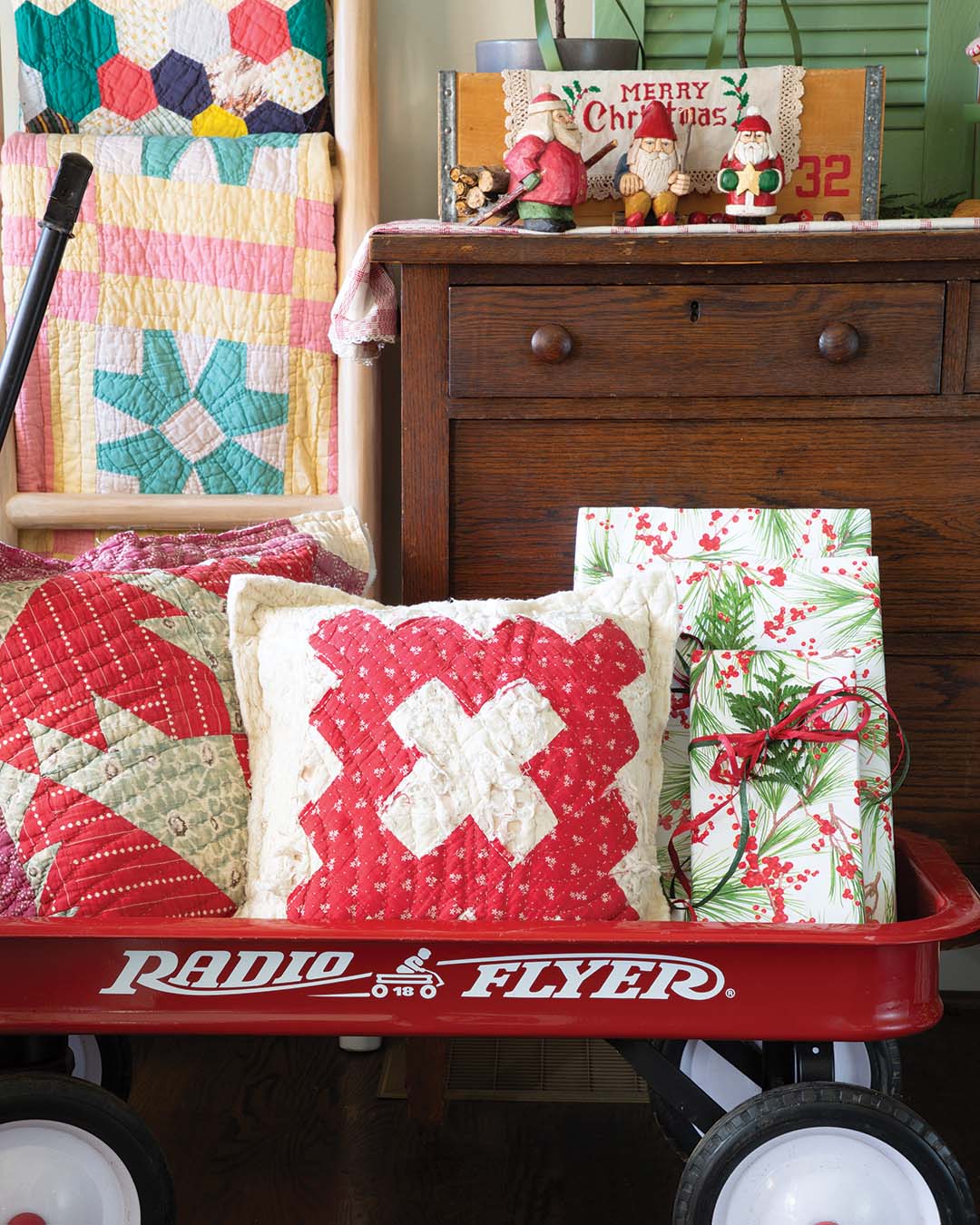 Colorful patchwork quilts displayed on a wooden ladder lend a dash of color and handmade artistry to the scene. Small wooden Santa figurines and a wreath accentuated with a cranberry-colored ribbon create an inviting vignette with a nostalgic note.