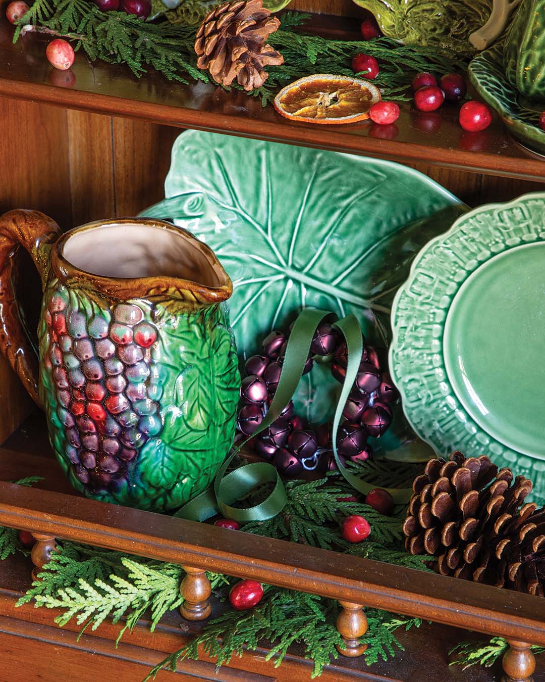 Fresh greenery drapes across warm wooden furniture enhanced by majolica dinnerware in various verdant designs, embracing the beauty of the outdoors. Bright red berry sprigs and pine cones line the hutch, adorning the space with the spirit of nature.