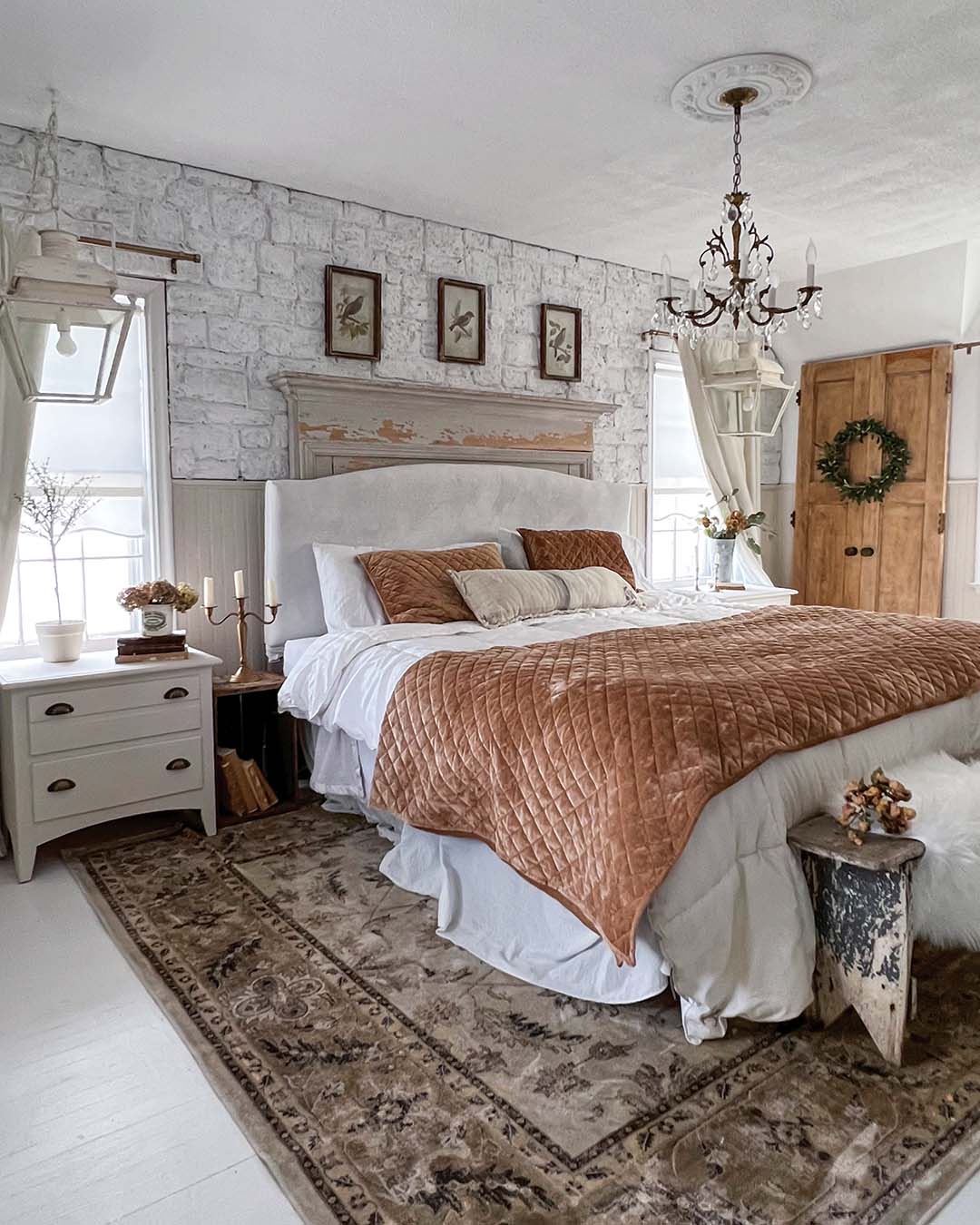 The primary bedroom continues Mickie’s go-to mingling of old and new with a striking faux-stone accent wall, a weathered mantel repurposed as a shelf above the headboard, large vintage-look lanterns that flank the bed, and an antique brass chandelier.