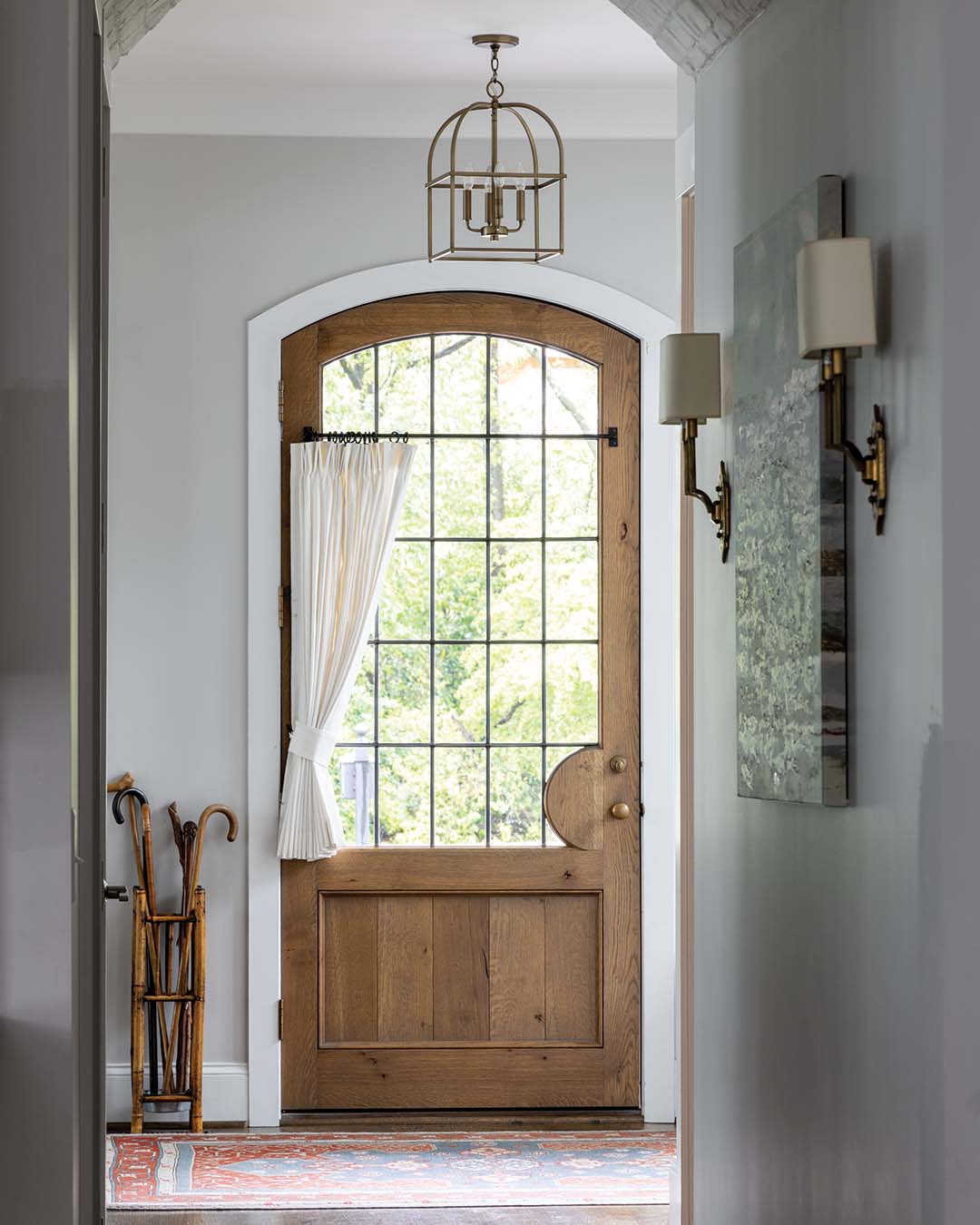 Warm wood tones and vibrant antique rugs set the tone for the home, beginning the moment you step inside. An arched wooden door stained in a honeyed hue draws the eye to the small hallway, beckoning guests further inside. An ivory café curtain softens the space, while the antique rug echoes the living room’s red-and-blue scheme.