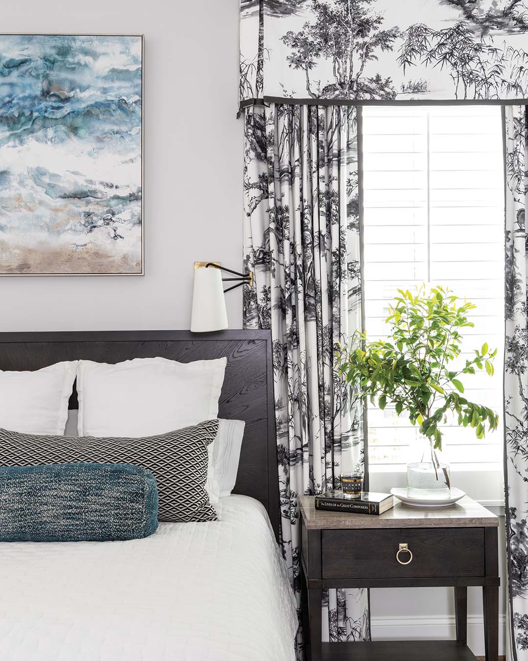 The primary bedroom draws inspiration from the abstract artwork hung above the bed. White quilted linens provide a soothing backdrop for the lumbar and bolster pillows to impart eye-catching contrast. Newlands Toile by Anna French dresses up the windows, providing a traditional final flourish.